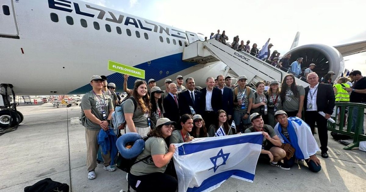 Groupe de jeunes juifs souriant brandissant des drapeaux israéliens sur le tarmac, devant un avion El Al.