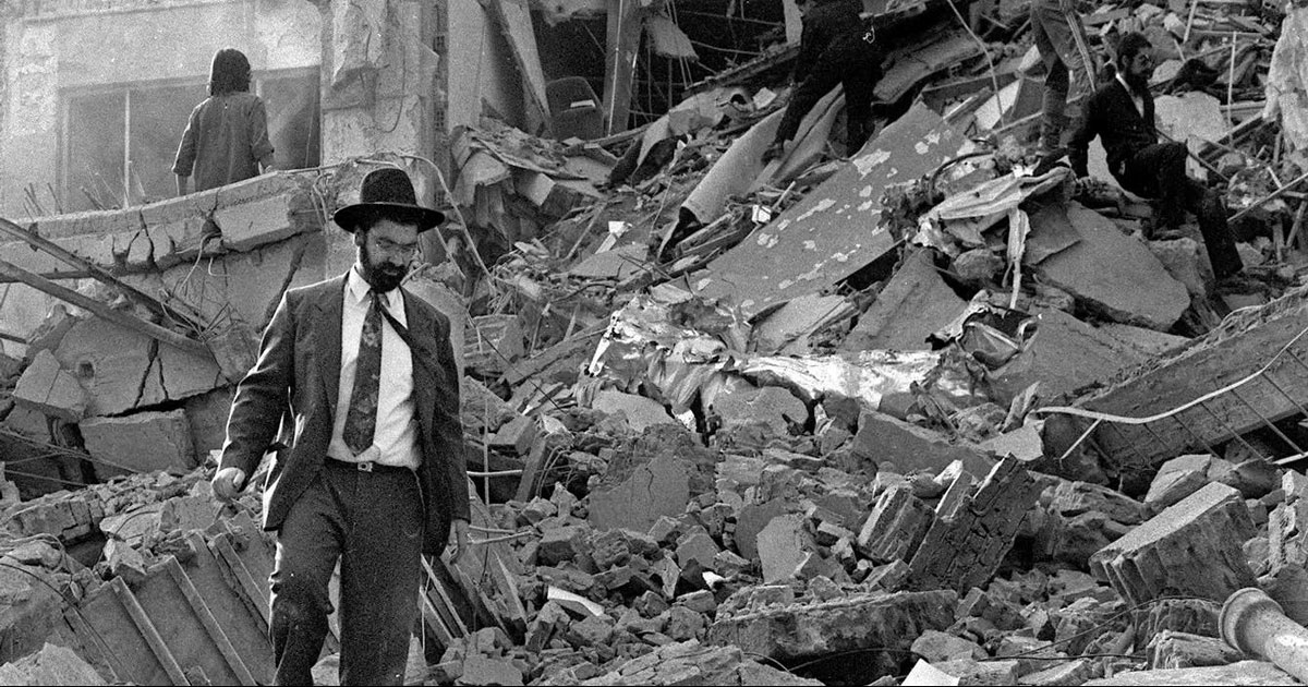Photographie en noir et blanc de la scène de l'attentat contre l'AMIA à Buenos Aires en 1994, montrant des sauveteurs et un membre de la communauté juive au milieu des décombres.