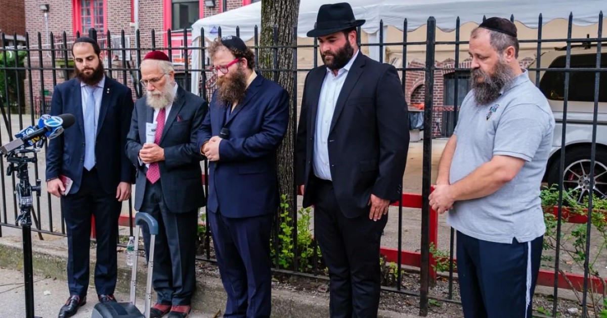 Cinq hommes juifs orthodoxes et 'hassidiques observent une minute de silence derrière des microphones lors d'une cérémonie commémorative en plein air à Brooklyn.