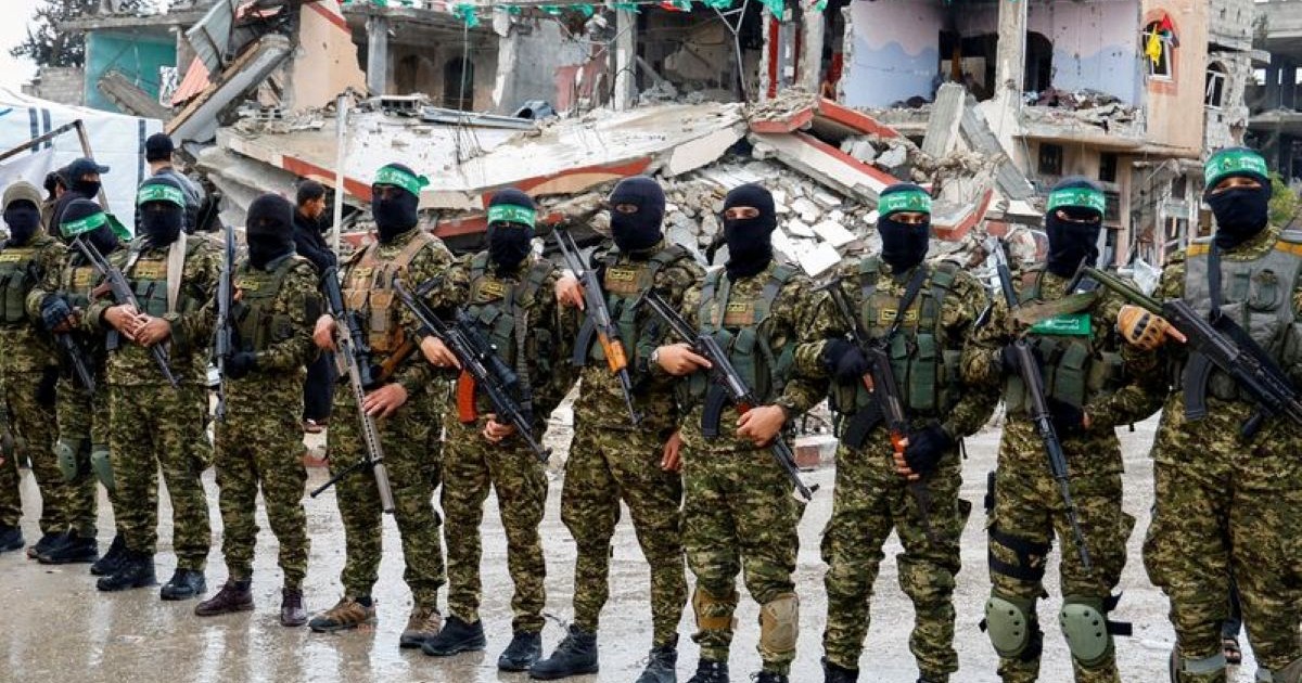 Un groupe d'hommes en uniforme de camouflage et cagoulés, armés de fusils, se tenant en rang devant un bâtiment détruit.