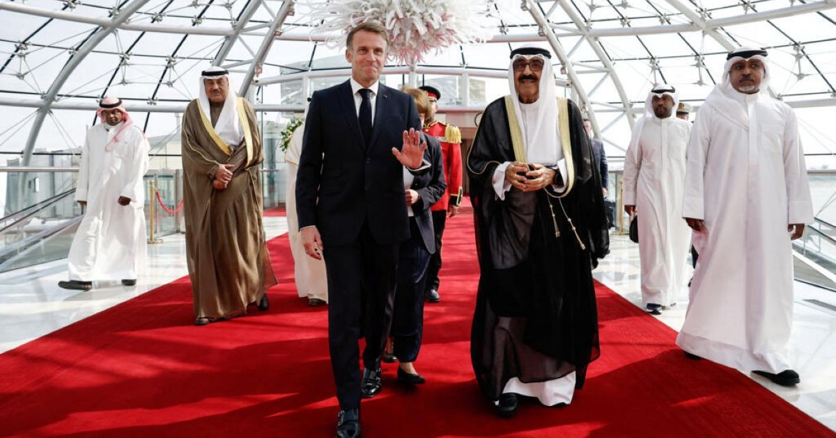 Emmanuel Macron salue de la main sur un tapis rouge, entouré de responsables koweïtiens sous une verrière.