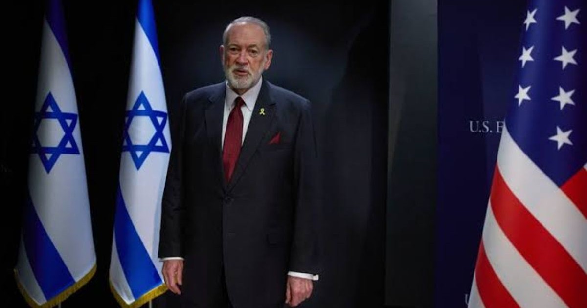 Mike Huckabee, ambassadeur américain en Israël, debout en costume devant les drapeaux israélien et américain.